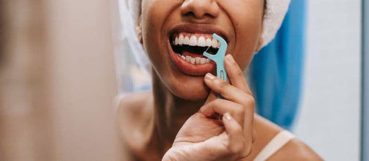 Femme souriante utilisant un porte-fil dentaire bleu pour nettoyer entre ses dents devant un miroir, illustrant la bonne technique d’hygiène bucco-dentaire.