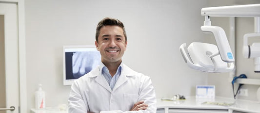 Chirurgien-dentiste souriant, bras croisés, dans un cabinet dentaire moderne équipé d’un fauteuil et d’un écran de radiologie, illustrant l’accompagnement professionnel pour les examens bucco-dentaires annuels.